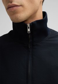 Schwarze Zip-Jacke mit Stehkragen, aus strukturiertem Stoff, sichtbarem Reißverschluss und kontrastierender innerer Kragenverzierung.