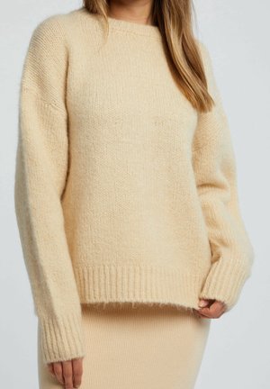 Femme portant un pull en maille doux beige à manches longues avec des poignets et un ourlet côtelés, assorti à une jupe côtelée.