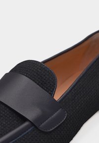 Κοντινή λήψη ενός σκούρου ναυτικού πλεκτού μοκασίνι με λείο δερμάτινο λουράκι και ταμπά εσωτερική επένδυση σε λευκό φόντο.