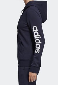 Sweat à capuche Adidas bleu marine avec logo blanc sur la manche, poche kangourou, capuche avec cordon ajustable et poignets et ourlet côtelés.