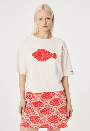 Frau mit langen blonden Haaren trägt ein weißes kurzärmeliges Oberteil mit rotem gehäkeltem Fisch und passendem roten Rock mit Fischmuster, stehend vor weißem Hintergrund.