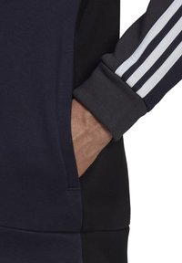 Sweat-shirt bleu marine avec une poche latérale, des poignets côtelés et trois bandes blanches sur les manches. Le matériau semble doux et texturé.