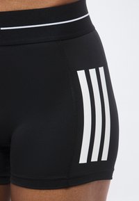 Gros plan sur un short noir moulant avec trois bandes blanches verticales sur le côté et une ceinture noire avec une fine ligne blanche.