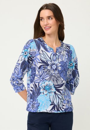 Woman with long brown hair wearing a blue and purple floral patterned blouse with three-quarter sleeves, smiling and looking to her left.