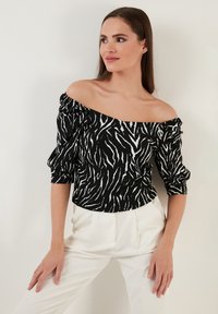 Černobílý off-the-shoulder top se zebřími pruhy, s nadýchanými rukávy, strukturovanou tkaninou a přiléhavým střihem. Nosený s bílými kalhotami.