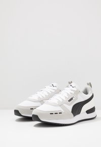 Valkoiset urheilujalkineet, joissa on harmaat mokkaakcentit ja musta sivujuova. Jalkineissa on verkkokangasta, tasaiset nauhat ja pehmustettu pohja.