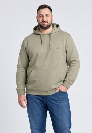 Man met baard die een licht olijfgroene hoodie en blauwe jeans draagt, staat met één hand in de zak tegen een lichte achtergrond.