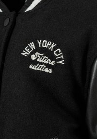 Mustmustest mustraha, millel on nahkvarrukad, valge triibuline krae ja tikandiga "New York City Tuleviku väljaanne" tekst ees.