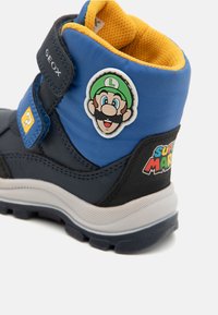 Baskets bleues et noires avec une sangle Velcro, arborant un badge du personnage Luigi et le logo "Super Mario" dans des couleurs vives. Tissu texturé et semelle en caoutchouc.