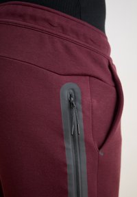 Maroon trenirke s glatkom teksturom, sa sivoj ziperskom džepom sa strane i udobnim pojasom.