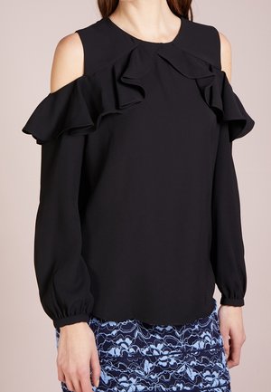Vrouw die een zwarte blouse met open schouders en ruches over de borst draagt, met lange mouwen, gecombineerd met een blauwe rok met bloemenpatroon.