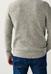 Homme portant un pull en maille gris texturé et un jean bleu foncé, vu de dos sur un fond clair.
