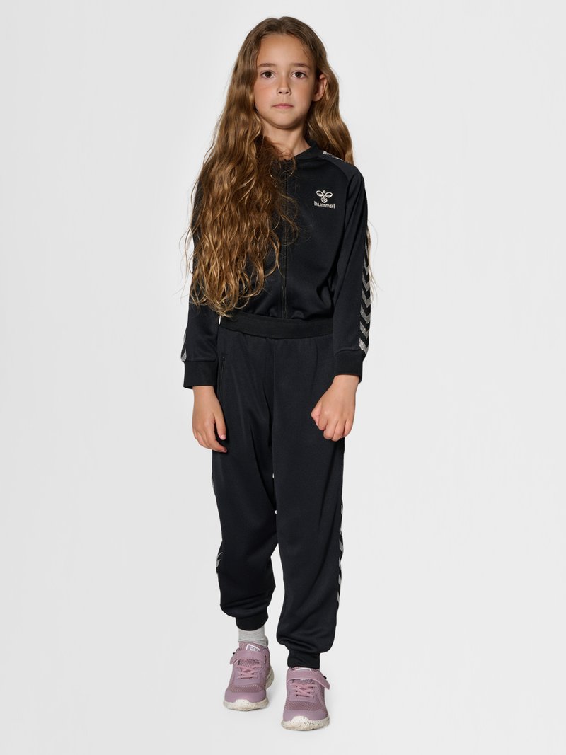 Jong meisje met lang haar, gekleed in een zwarte jas met rits en broek met chevronstrepen, staand tegen een effen lichte achtergrond.