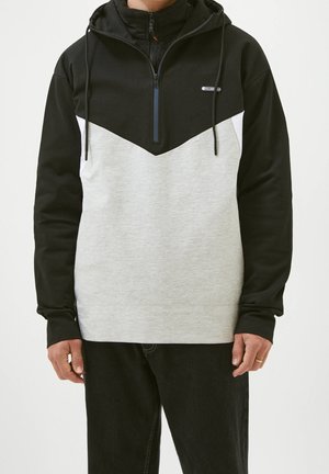 Homme portant un sweat à capuche noir et gris avec une demi-fermeture éclair et des cordons de serrage, assorti d'un pantalon noir, se tenant devant un fond uni.
