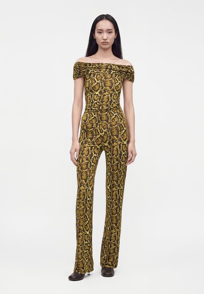 Γυναίκα με μακριά μαύρα μαλλιά, φοράει ένα κίτρινο και καφέ ολόσωμο φόρεμα με σχέδιο φίδι και χωρίς ώμους, καθώς και μαύρα παπούτσια, στέκεται σε λευκό φόντο.