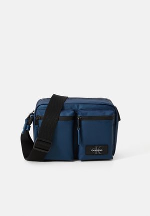 Borsa a tracolla blu navy con due tasche frontali con cerniera, una tracolla nera regolabile e una patch con logo. Realizzata in materiale liscio e resistente.
