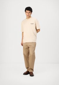 Beige cotton t-shirt with short sleeves and black logo on the chest, paired with light brown trousers and dark brown loafers.