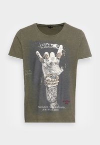 T-shirt en coton vert olive avec un graphisme d'une main faisant un geste de rock, accompagné de texte et de tatouages détaillés. Encolure ronde, manches courtes.