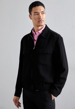 Chaqueta negra texturizada con dos bolsillos en el pecho y cremallera frontal. Llevada sobre una camisa rosa claro con lunares rojos.