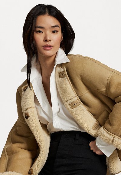 Giacca in shearling marrone con dettagli in bottoni, camicia bianca con colletto sotto, e pantaloni neri. Il tessuto presenta una finitura testurizzata.