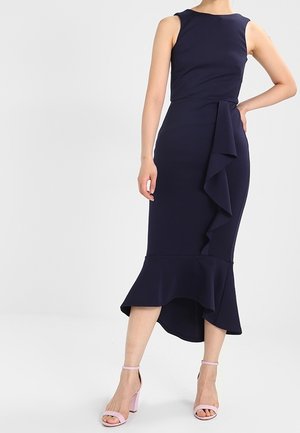 Mouwloze navy midi-jurk met asymmetrische ruches aan de voorkant en zoom, gecombineerd met lichtroze sandaaltjes met blokhak en enkelriem.