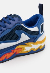 Baskets bleu et blanc avec un mesh bleu foncé, fermeture à lacets à l'avant et motif de flammes jaunes et oranges sur le côté de la semelle, avec une languette au talon.