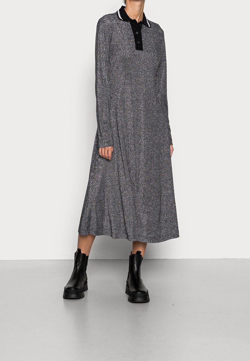 Femme portant une robe midi argent-grise à manches longues avec un col noir et des bottines noires, se tenant contre un mur blanc uni.