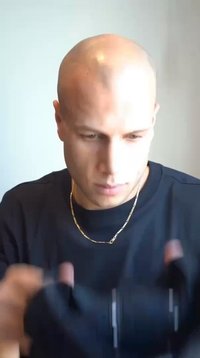 Vyras juoda apvalios iškirptės marškinėliais, koncentruojasi į kamerą. Jis plikas ir dėvi ploną auksinę grandinėlę ant kaklo.