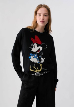Junge Frau trägt einen schwarzen Pullover mit einem großen Minnie-Maus-Motiv in Rot, Blau, Weiß und Gelb, posiert mit Händen in den Taschen vor weißem Hintergrund.