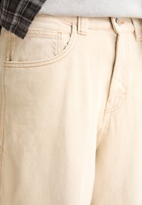 Pantalon en denim beige clair avec poche avant visible et passants pour ceinture, associé à une chemise à carreaux rentrée et un haut gris.