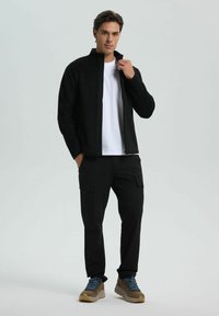 Černá zipová bunda přes bílý tričko, v kombinaci s černými cargo kalhotami. Hnědé turistické boty s modrými tkaničkami. Hladká textura, ležérní design.