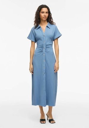 Žena v modrom midi šatách na gombíky s krátkymi rukávmi a riaseným pásom, v kombinácii s čiernymi otvorenými lodičkami na podpätku, stojaca na bielom pozadí.