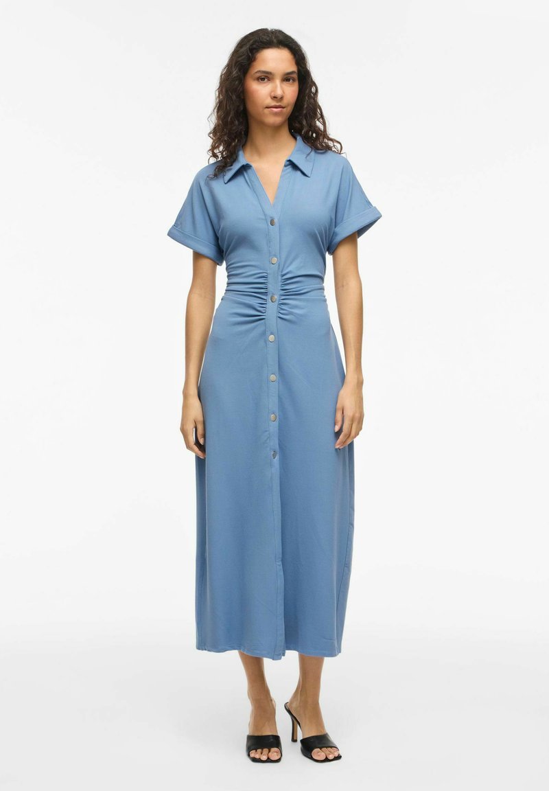 Femme portant une robe midi bleue boutonnée, manches courtes et taille froncée, associée à des mules à talons noires à bout ouvert, debout sur fond blanc.