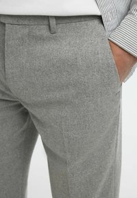 Pantaloni grigi in lana con una finitura testurizzata, dotati di una tasca singola e un design a vita piatta, abbinati a un polsino di camicia a righe.