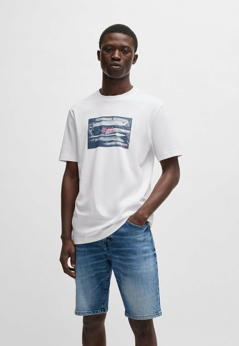 T-shirt bianca in cotone con stampa grafica raffigurante mani e testo. Abbinata a pantaloni shorts in denim azzurro chiaro, che mostrano un fit casual.