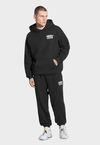 Czarny zestaw z kapturkiem i dresowymi spodniami wykonany z materiału mieszanki bawełny, z przednią kieszenią oraz białymi detalami logo na klatce piersiowej i udzie.