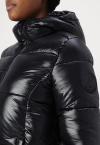 Zwarte puffy jas met een glanzende afwerking, voorzien van een ritssluiting aan de voorkant, een capuchon en een ronde logopatch op de linkerschouder. Gelaagd quiltontwerp.