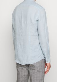 Uomo che indossa una camicia a maniche lunghe azzurro chiaro e pantaloni a quadri grigi, visto da dietro su uno sfondo neutro.
