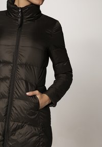 Svart puffjacka med hög krage, dragkedja framtill och vertikal quiltad design. Har sidofickor och en slät, glänsande yta.