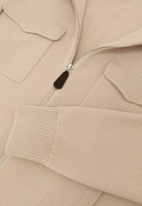 Bēša rāvējslēdzēja džemperis ar ribojumiem piedurknēs un apkaklē, divām kabatām uzšuvumos un gludu tekstūru ar tīru apdari.