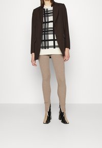 Blazer marron ajusté porté sur un pull à carreaux noir et blanc, associé à des leggings côtelés beige et des bottines en cuir verni noir avec un accent beige.