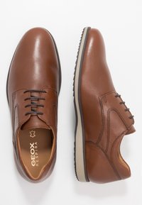 Chaussures en cuir marron avec une finition lisse, bout rond, fermeture à lacets à six œillets, et détails texturés sur les côtés. Semelle contrastée.