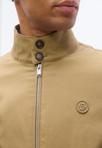 Άνδρας που φοράει καφέ μπουφάν με ψηλό γιακά, δύο μαύρα κουμπιά, ορατό ασημένιο φερμουάρ και μικρό κεντημένο λογότυπο στο στήθος.