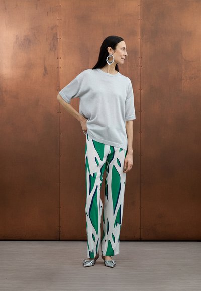 Haut ample gris clair à manches courtes associé à un pantalon blanc orné de motifs abstraits verts et bleus. Chaussures plates argentées et boucles d'oreilles.