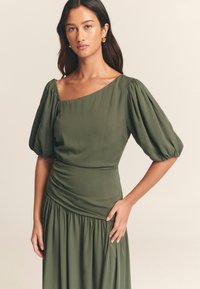 Abito verde oliva con maniche a sbuffo, scollatura asimmetrica, corpetto aderente e vita arricciata che si allarga in una gonna plissettata. Tessuto morbido.