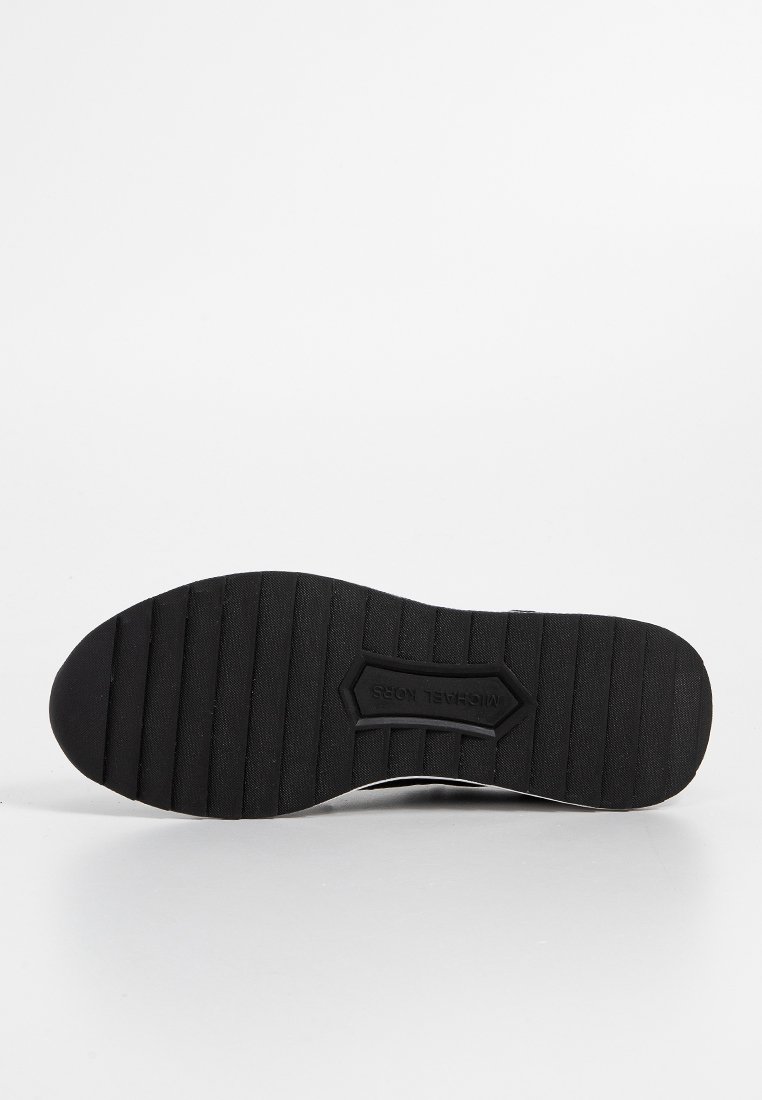 Suola di scarpa in gomma nera con texture rigata, caratterizzata da un'etichetta con logo in rilievo. Bordo liscio e design della punta leggermente appuntita.