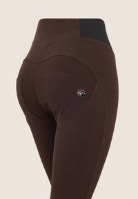 Leggings marroni con vita alta, cuciture curve e dettagli sui taschini. Il materiale appare liscio con una leggera lucentezza.