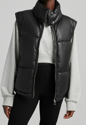 Gilet rembourré sans manches en simili cuir noir avec col montant, fermeture éclair et boutons-pression. Porté par-dessus un sweat-shirt court gris clair.