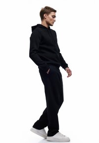 Sort hættetrøje og matchende sweatpants, lavet af blødt stof, med en afslappet pasform. Modellen bærer hvide sneakers og står i profil.