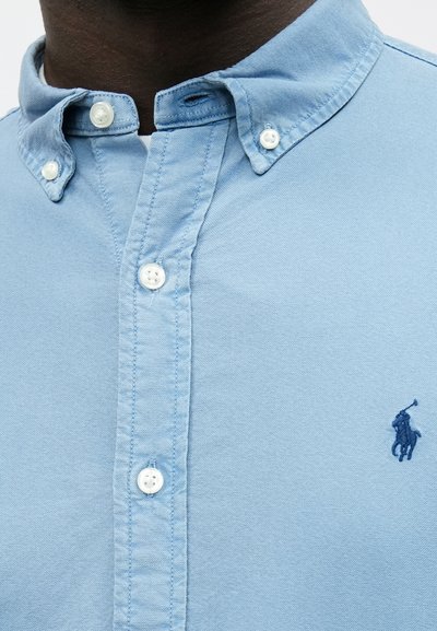 Camisa de algodão azul claro com botões, gola clássica, botões brancos e pequeno logótipo azul-marinho no lado esquerdo.